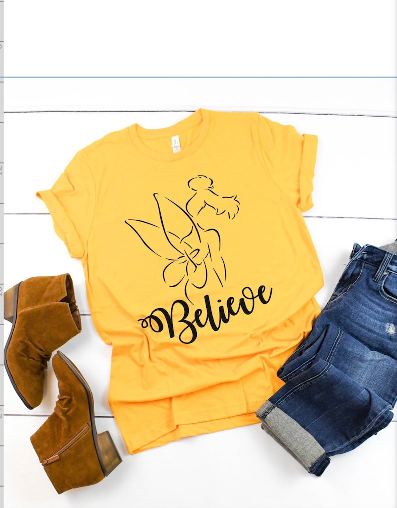Puede incluir: Una camiseta amarilla con un contorno negro de Campanilla y la palabra "Believe" en letra negra.