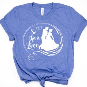 May include: A blue t-shirt featuring a white graphic of Cinderella and Prince Charming dancing. The graphic is within a circle with the text "So this is Love" written in a cursive font.