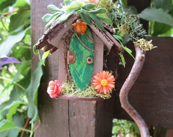 May include: A miniature fairy house with a green door, surrounded by moss, flowers, and greenery. The house is made of wood and has a brown roof.