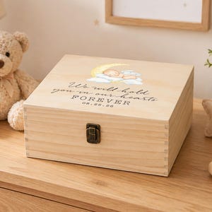 May include: A rectangular light-coloured wooden keepsake box with a metal latch. The lid features a watercolour illustration of a baby sleeping on a crescent moon and clouds, with the text "We will hold you in our hearts FOREVER 08.22.26".