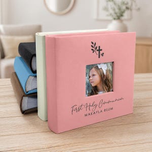 May include: A stack of photo albums in various colors, including pink, black, blue, and brown. The front album is pink with a photo cutout and the text "First Holy Communion" and a name. A cross and leaf design is above the photo.