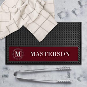 May include: A black bar mat with a textured surface and a burgundy nameplate that reads "MASTERSON" in white. A white and gray checkered towel and ice tongs are also present. Ice cubes are scattered around the mat.