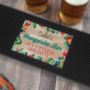 May include: A black bar mat with a colorful rectangular design that reads "Caleb's Margarita Bar" with the phrase "It's 5 O'Clock Somewhere". The design features parrots, tropical flowers, and foliage.