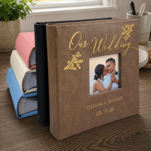 May include: A square brown photo album with a textured cover featuring gold foil text that reads Our Wedding, Olivia + Wayne, and 05.15.25. A square cutout displays a photograph of a couple. Behind it are three additional albums in black, cream, and blue.
