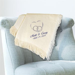 May include: Cream-colored throw blanket with a fringed edge, embroidered with a heart and two intertwined rings. The blanket has the names "Matt & Casey" and the date "April 19, 2025" embroidered in navy blue.