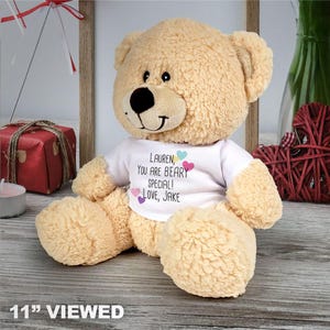 May include: A light tan plush teddy bear wearing a white t-shirt. The shirt features the text "Lauren, you are beary special! Love, Jake" printed in black, accented with small pink, blue, and purple heart icons. The bear has dark eyes and a nose.