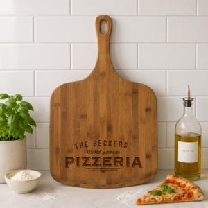 May include: A wooden pizza peel with a handle, engraved with "THE BECKERS World Famous PIZZERIA." A slice of pizza, a bottle of olive oil, a small bowl of flour, and a potted basil plant are also visible.