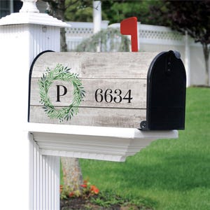 May include: A mailbox featuring a faux grey wood grain finish with a green leaf wreath graphic surrounding the letter P and the house number 6634 printed in black. The mailbox has a black metal front door and a bright red signal flag on top.
