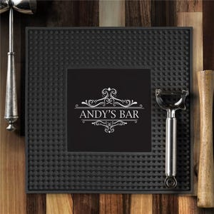 May include: A black bar mat with a textured surface and the text "ANDY'S BAR" in white. The mat is surrounded by bar tools, including a cocktail shaker, a peeler, and a muddler. The background is a wooden surface.