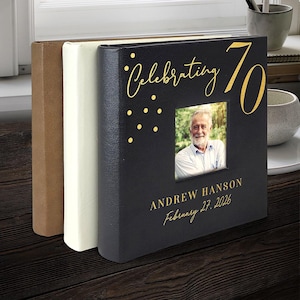 May include: A black photo album with gold lettering that reads "Celebrating 70". The album has a photo of a man and the name "Andrew Hanson" and the date "February 27, 2026". Two other albums are visible.