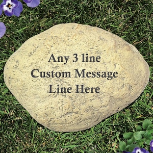 May include: A tan, oval-shaped garden stone with a textured, rock-like surface. The center of the stone features dark, engraved text that reads: Any 3 line Custom Message Line Here. The stone has an irregular, natural edge and a flat top surface.