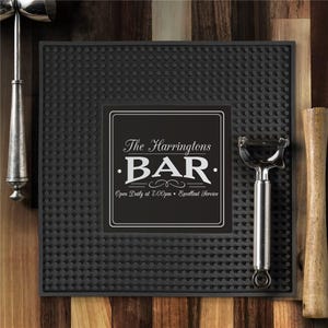 May include: A black bar mat with a textured surface and a rectangular label. The label reads "The Harringtons BAR" in white text. A cocktail shaker, a peeler, and a muddler are on the sides.
