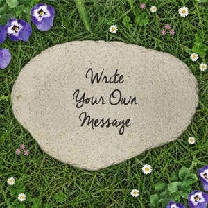 Peut inclure: Une pierre de jardin ovale de couleur gris clair avec une surface texturée et tachetée. Le texte "Write Your Own Message" est imprimé en lettres cursives noires au centre de la pierre, qui est posée sur de l'herbe verte.