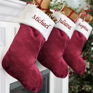 Peut inclure: Trois chaussettes de Noël en velours bordeaux profond avec des revers en peluche blanche. Chaque revers est brodé d'un prénom en écriture rouge : Michael, Juliana et Sophia. Les chaussettes contiennent des paquets en papier brun et des sucres d'orge.