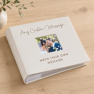 May include: A square, off-white photo album with a central square cutout displaying a photograph of a couple in wedding attire. The cover features the printed text "Any Custom Message" above the photo and "WRITE YOUR OWN MESSAGE" below it.