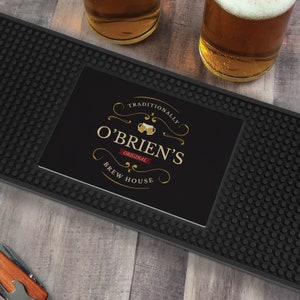 May include: A rectangular black rubber bar mat with a textured surface featuring a central graphic with the text "TRADITIONALLY O'BRIEN'S ORIGINAL BREW HOUSE" in gold and white lettering, accented by two small beer mug icons and gold swirls.