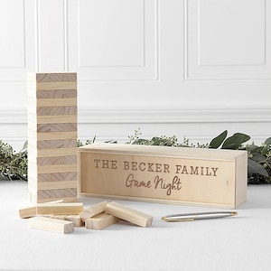 May include: A light-colored wooden block stacking game with a rectangular storage box featuring the engraved text THE BECKER FAMILY Game Night. Several loose wooden blocks are scattered in front of the box, and a silver pen lies nearby.