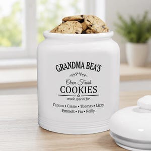 May include: A white ceramic cookie jar with a lid, featuring black text that reads GRANDMA BEA'S Oven Fresh COOKIES made special for Carson, Cassie, Thomas, Lizzy, Emmett, Fin, and Reilly. The jar is filled with several chocolate chip cookies.