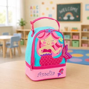 May include: A bright blue and pink lunch bag featuring a cartoon mermaid with pink hair, a crown, and a star wand. The lower pink section displays the name Amelia in dark purple script, accompanied by two small seashell illustrations on the side.
