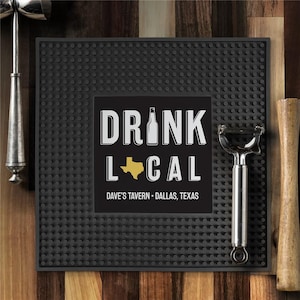 May include: A black bar mat with a textured surface and the words "DRINK LOCAL" in white, with a gold Texas outline and the text "DAVE'S TAVERN - DALLAS, TEXAS". A cocktail shaker, peeler, and muddler are on the sides.