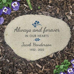 May include: A light beige, oval-shaped memorial stone with a textured surface. The stone features blue floral accents and the text: Always and forever IN OUR HEARTS, Jacob Henderson, 1932 - 2022. The lettering is dark gray and clearly legible.