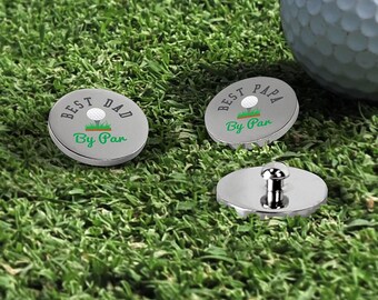 May include: Two circular silver metal golf ball markers featuring a graphic of a white golf ball on a green tee. One marker displays the text BEST DAD By Par, and the other displays BEST PAPA By Par. A third marker shows the silver metal post underside.