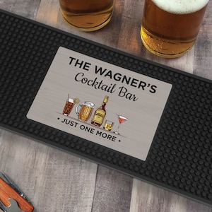 May include: A rectangular black rubber bar mat with a textured surface and a central silver-toned insert. The insert features the text THE WAGNER'S Cocktail Bar and JUST ONE MORE, accompanied by illustrations of a mixed drink, beer mug, whiskey bottle, glass, and martini glass.