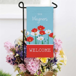 May include: A garden flag with a light blue background, featuring the name "The Wagners" and the word "WELCOME" in white. The flag has red and white flower illustrations. A black metal garden stake holds the flag, with a bouquet of flowers in the foreground.