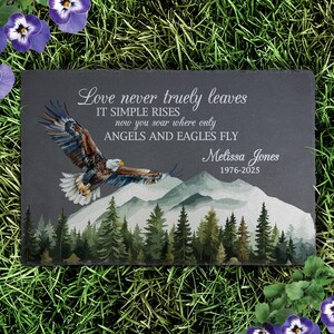 May include: A rectangular dark gray slate memorial plaque featuring a printed graphic of a bald eagle in flight over a mountain range and pine forest. The text reads: Love never truly leaves IT SIMPLE RISES now you soar where only ANGELS AND EAGLES FLY Melissa Jones 1976-2025.