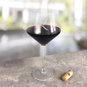 May include: A clear glass wine glass filled with deep red wine, featuring a white etched letter L on the bowl. The stem is slender and connects to a circular base. A small, cylindrical natural cork is positioned to the right of the glass.