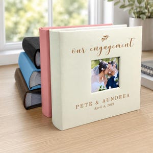 May include: A square cream-colored photo album with a central window displaying a couple. The cover features gold text reading our engagement, PETE & AUNDREA, and April 4, 2025. Behind it are stacked albums in pink, black, blue, and brown colors.