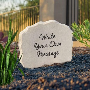 May include: A light-colored, irregular, stone-like garden plaque with a rough, textured edge. The center of the plaque features the black, cursive text "Write Your Own Message" printed clearly on the smooth, flat surface of the stone.