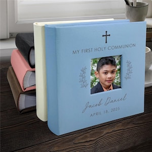 May include: A stack of photo albums in various colors, including blue, pink, and black. The front album is light blue with a cross and the text "MY FIRST HOLY COMMUNION." It features a photo of a child and the name "Jacob Daniel" with the date "APRIL 18, 2025."