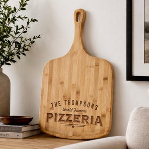 May include: A light brown bamboo pizza peel with a long handle and a hole for hanging. The surface features dark engraved text that reads: THE THOMPSONS World Famous PIZZERIA with a small star symbol centered below the word PIZZERIA.