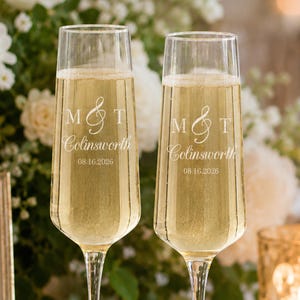 May include: Two clear glass champagne flutes filled with a sparkling golden liquid. Each flute is etched with the letters M and T separated by an ampersand, the name Colinsworth, and the date 08.16.2026 in a white, elegant serif font.