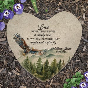 May include: A heart-shaped stone memorial plaque featuring a printed illustration of a bald eagle flying over a forest and mountains. The text reads: Love NEVER TRULY LEAVES it simply rises. NOW YOU SOAR WHERE ONLY angels and eagles fly Melissa Jones 1976-2025.