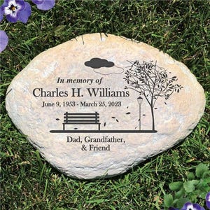May include: A light-colored, oval-shaped memorial stone featuring black engraved text that reads In memory of Charles H. Williams, June 9, 1953 - March 25, 2023, and Dad, Grandfather, & Friend, alongside a graphic of a tree, a bench, and a cloud.