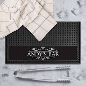 May include: A black bar mat with the text "ANDY'S BAR" in white script. A white and gray checkered bar towel rests on the mat. Stainless steel ice tongs and ice cubes are also visible.
