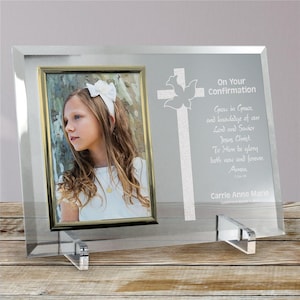 May include: A clear glass photo frame with a gold-trimmed photo of a young girl. The frame has a cross and dove design with the text "On Your Confirmation" and a prayer. The frame is supported by clear acrylic stands.
