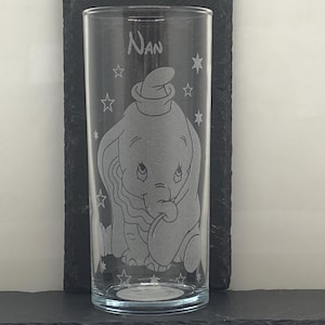 Puede incluir: Un vaso de vidrio transparente grabado con un diseño de Dumbo el elefante y el nombre "Nan". El diseño incluye estrellas alrededor del elefante. El vaso se muestra sobre una superficie oscura.