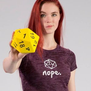 May include: A woman with long red hair is holding a yellow 20-sided die. She is wearing a purple shirt with a white graphic of a die and the word "nope."