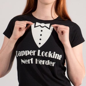 May include: A black t-shirt with a white tuxedo graphic and the text "Capper Looking Nerf Herder". The model is wearing blue jeans.
