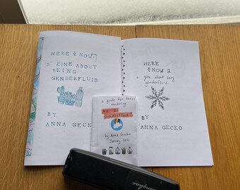 Può includere: Tre zine con copertine bianche e testo nero. Quello a sinistra riporta Here & Now: A Zine About Being Genderfluid di Anna Gecko con illustrazioni di cactus. Quello a destra riporta Here & Now 2 di Anna Gecko con un fiocco di neve.
