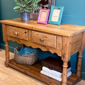 May include: A light brown wooden console table with two drawers and a lower shelf. A pink and a teal picture frame with the text "Paper High" sit on top. A wicker basket, books, and a small suitcase are on the shelf.