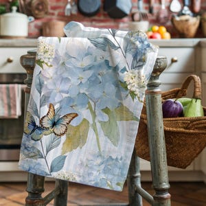 Può includere: Un canovaccio da cucina bianco con un motivo di ortensie blu e farfalle. Il canovaccio presenta un motivo floreale in stile acquerello con fiori azzurri e bianchi, foglie verdi e due farfalle. Il canovaccio è drappeggiato su uno sgabello di legno.