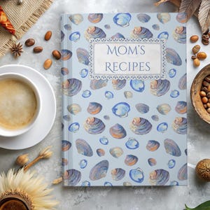 May include: A recipe book titled "Mom's Recipes" with a light blue cover adorned with a seashell pattern. The design includes blue, purple, and orange seashells. The title is framed in a decorative border.
