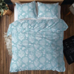 May include: A light blue duvet cover and two pillowcases with a white coral and seashell pattern. The bedding set is displayed on a bed with a wooden headboard and a wooden floor. The overall theme is coastal.