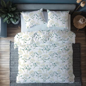 May include: White duvet cover and two pillowcases with a floral design. The pattern shows green stems and yellow flower clusters. The bedding is on a bed with a light blue sheet, a grey rug, and a wooden side table.