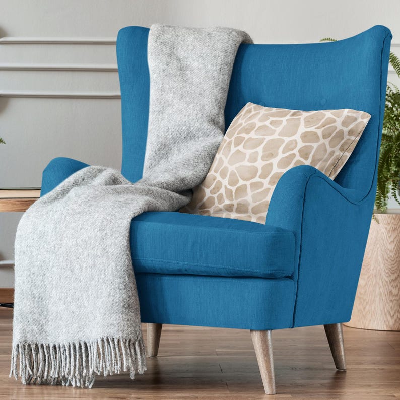 Pu&ograve; includere: Una poltrona blu con una coperta di lana grigio chiaro e un cuscino beige con un motivo geometrico. La poltrona ha gambe in legno e uno schienale imbottito.