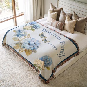 May include: A white throw blanket with a blue border and fringe. The blanket features a floral design with blue hydrangeas and butterflies. The text "Brayton Family EST 2026" is printed on the blanket.
